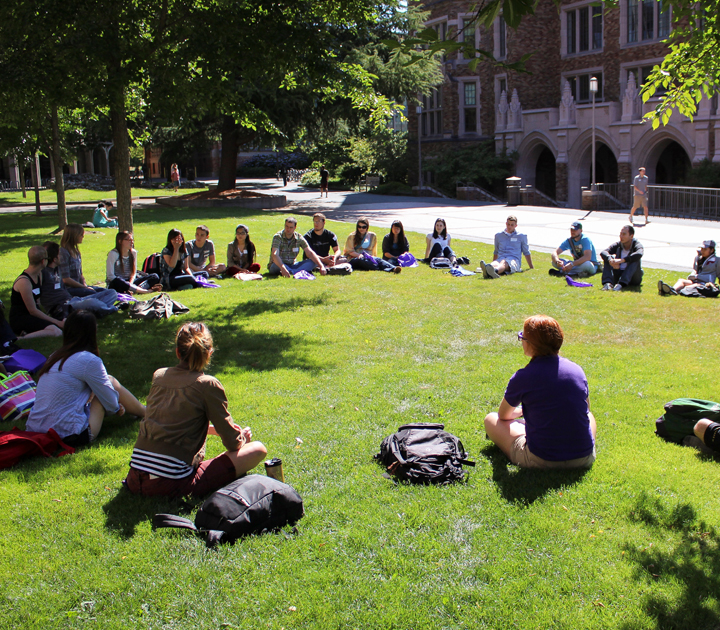 Circle of students sit outside on grass and have class.