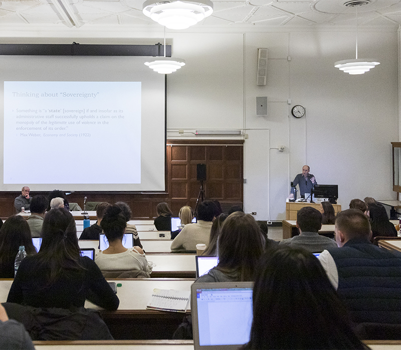 Teacher talks at podium in front of lecture hall with projection screen