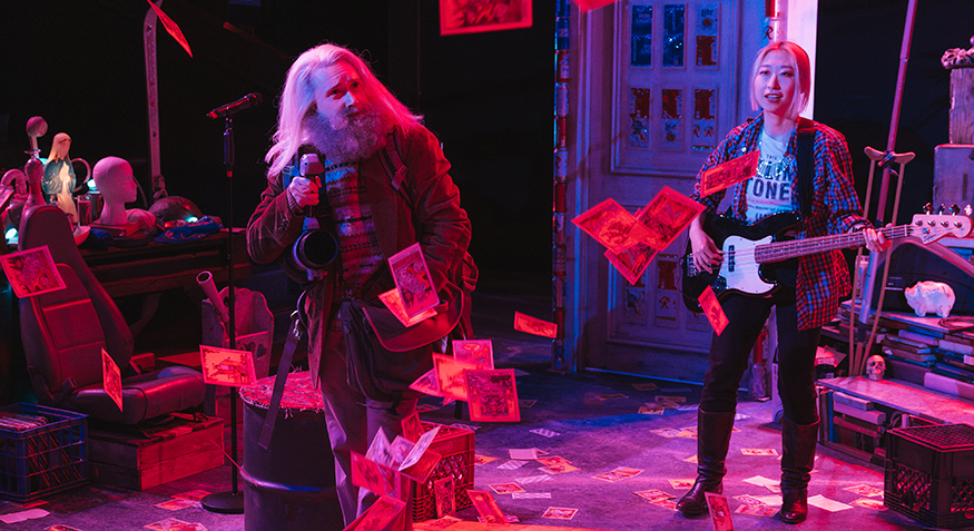 Performance with stage flooded in pink/purple lighting with tarot card flying through the airor playing guitar, the other holding a microphone, with paper bi